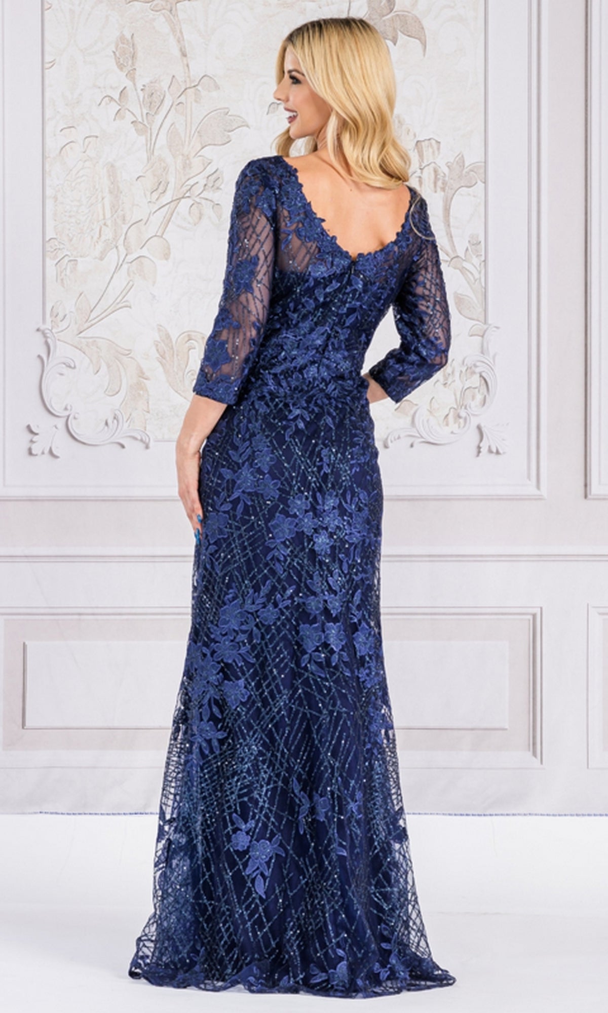 A woman with blonde hair models the Floral-Embroidered Long Sleeve Formal Dress 7045, a navy blue gown with delicate floral embroidery and long sleeves, posing in front of a decorative wall with her back to the camera.
