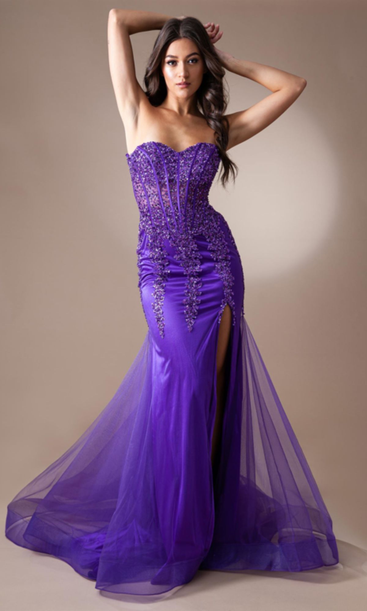 Model poses against a neutral background in the Strapless Long Beaded Sheer-Bodice Prom Dress 7051, featuring a corset-style bodice, floor-length purple skirt, beaded details, and a high slit.