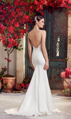 A woman in the Long White Dress: Alyce 7051 with a cowl neckline stands outdoors by a rustic green door, surrounded by red flowers and potted plants.
