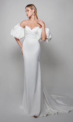 A woman wearing the Alyce 7056 long formal dress—white, off-the-shoulder with a sweetheart neckline, puffed sleeves, and fitted bodice—stands against a plain light background.
