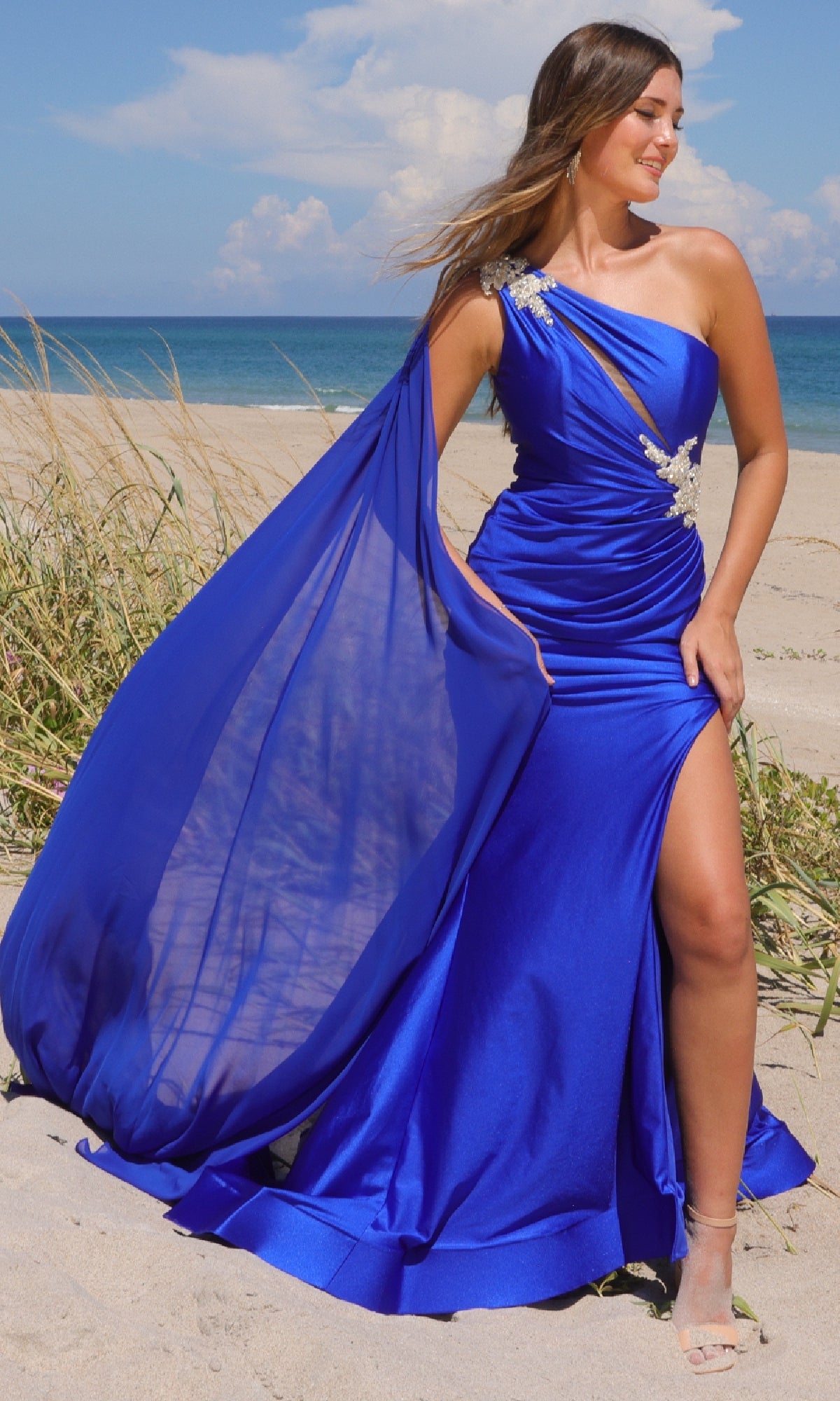 A woman wearing the Amelia Couture 7056 One-Shoulder Long Prom Dress, featuring a sheer cape and thigh-high slit, stands on a sandy beach with grass and ocean in the background.