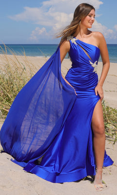 A woman wearing the Amelia Couture 7056 One-Shoulder Long Prom Dress, featuring a sheer cape and thigh-high slit, stands on a sandy beach with grass and ocean in the background.