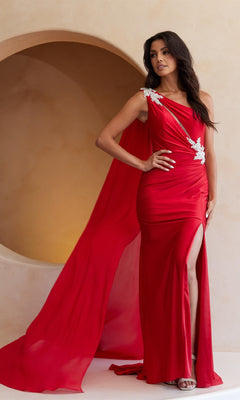 A woman in the Amelia Couture 7056, a bright red one-shoulder long prom dress with white floral details, flowing cape, and thigh-high slit, stands indoors against a curved beige backdrop.