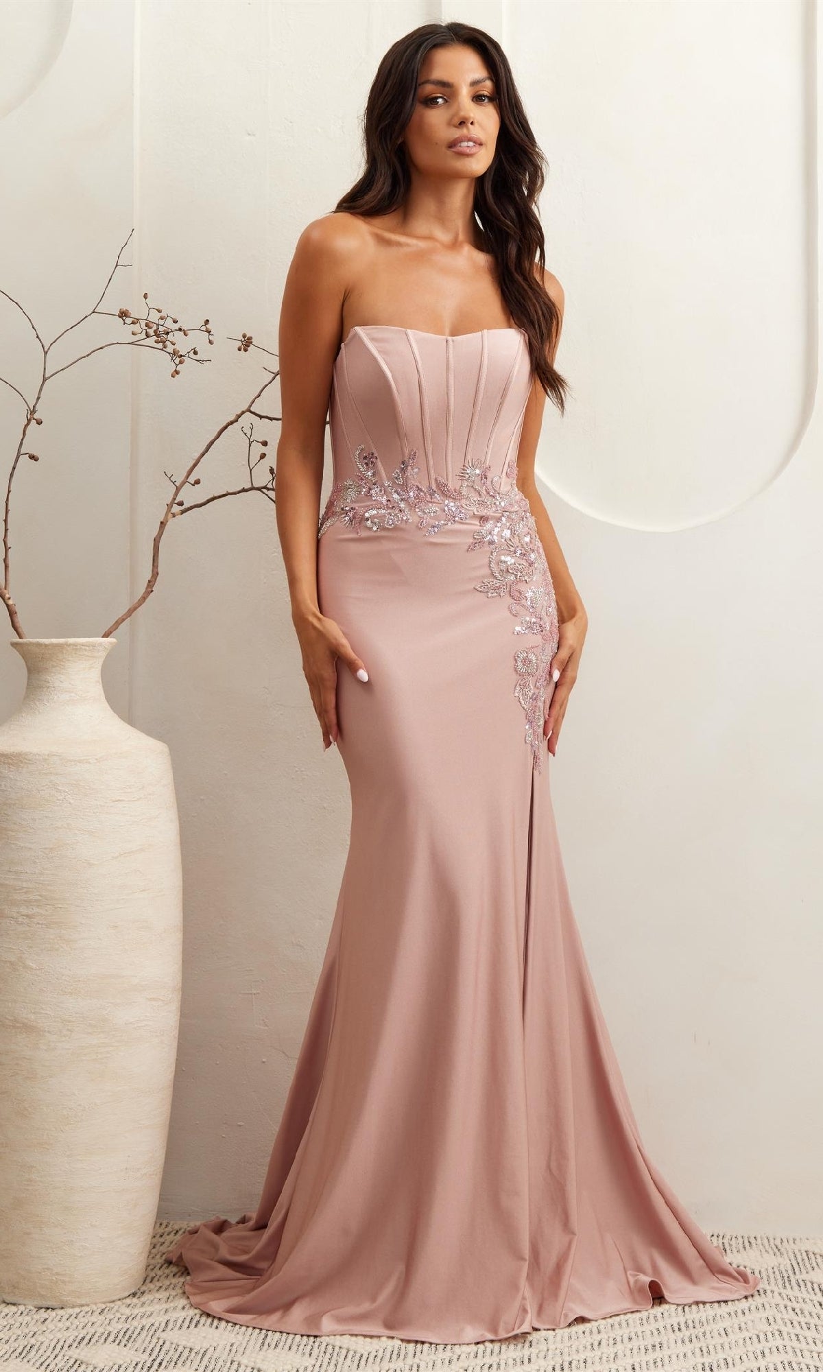 A woman in the Amelia Couture 7057 strapless corset prom dress with sequin lace appliques stands beside a tall vase with branches against a light background.