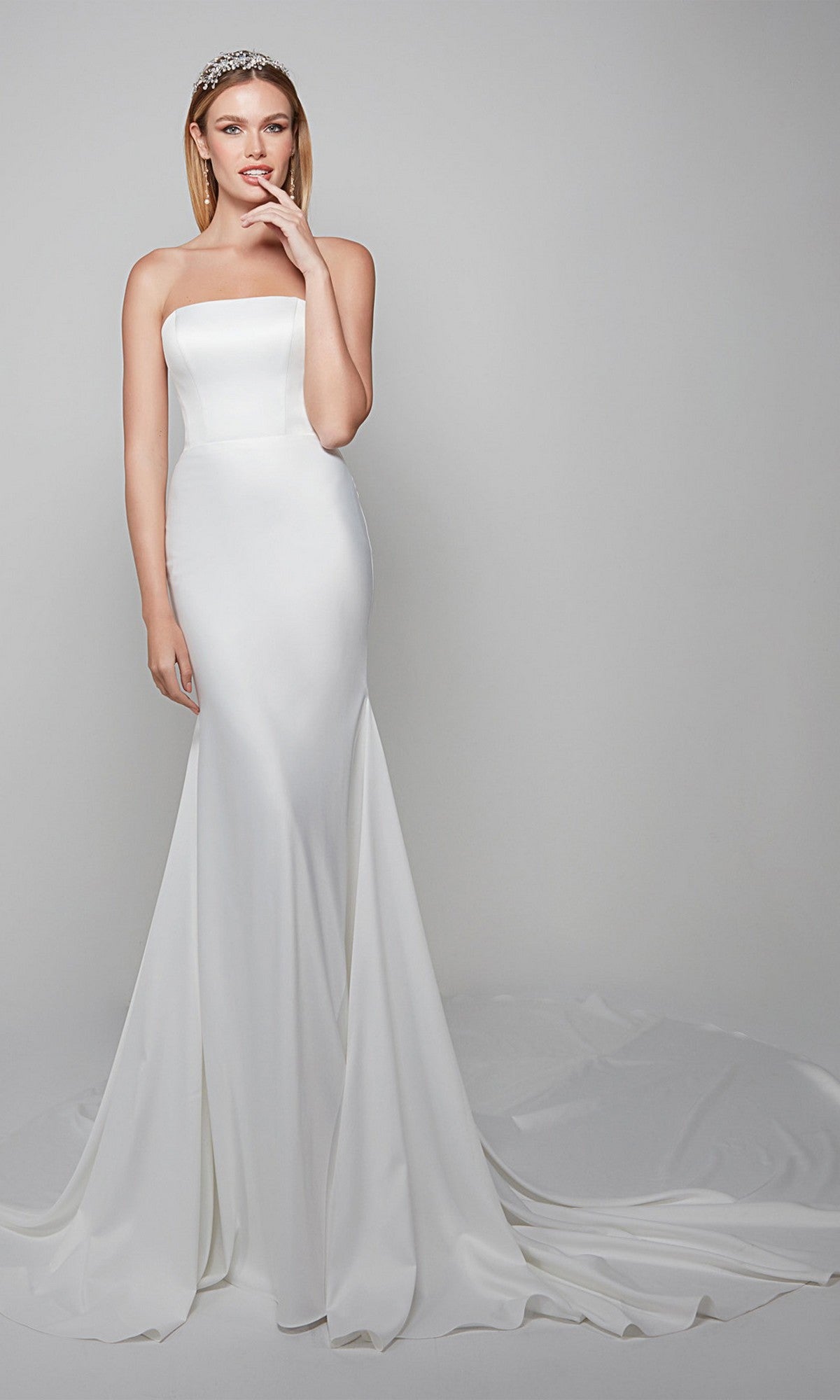 A woman in the Alyce 7060 long dress in diamond white, featuring a strapless fitted satin trumpet silhouette and long train, stands against a plain light background, touching her lips and wearing a jeweled headband.