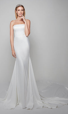 A woman in the Alyce 7060 long dress in diamond white, featuring a strapless fitted satin trumpet silhouette and long train, stands against a plain light background, touching her lips and wearing a jeweled headband.