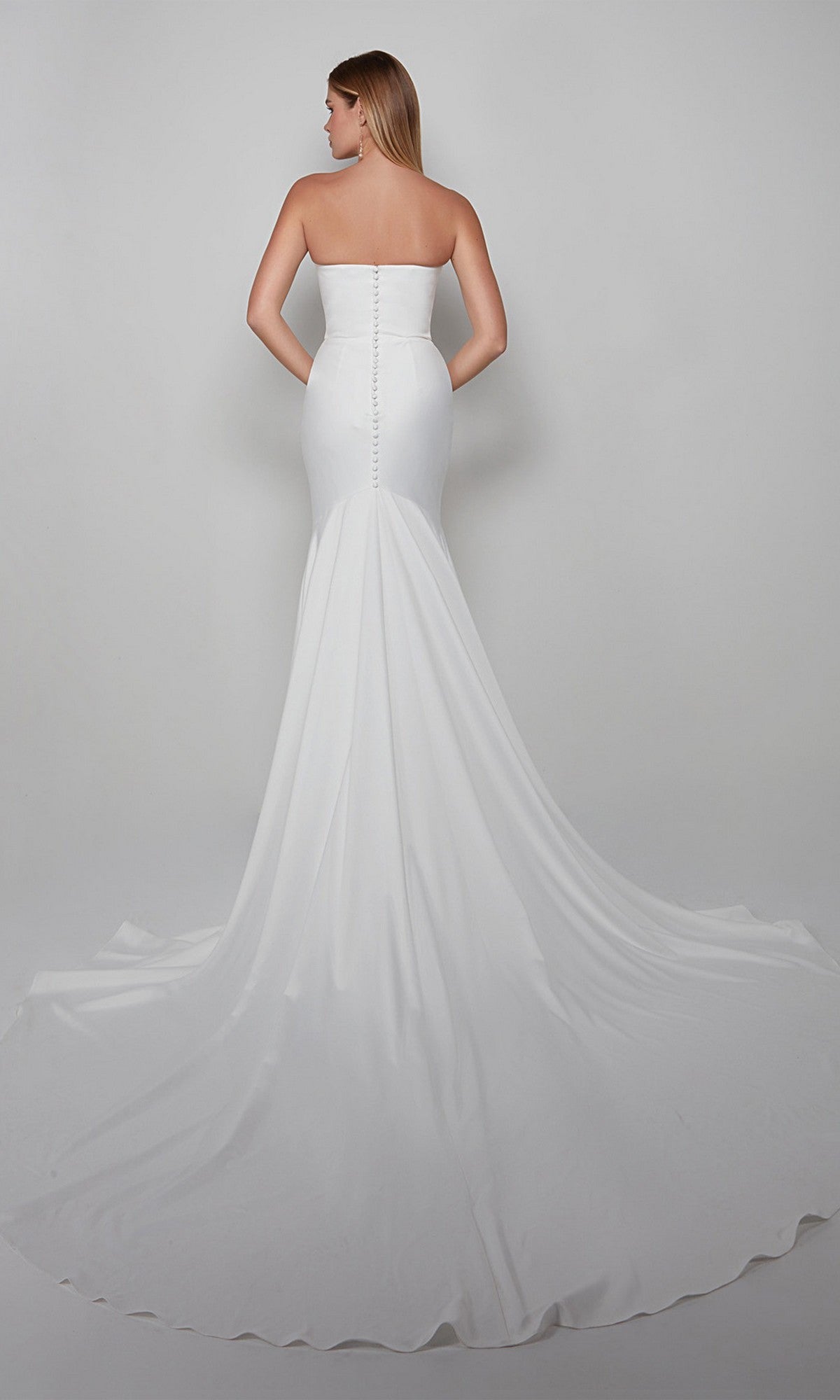 A woman is photographed from the back in a strapless, fitted satin Long White Dress: Alyce 7060 with a trumpet skirt and long train, set against a plain light gray background.