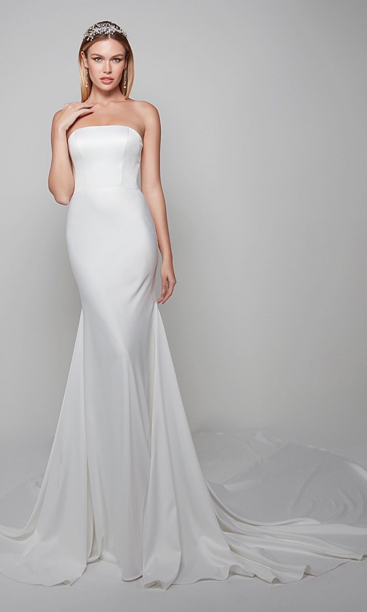 A woman poses against a plain light gray background, wearing the Alyce 7060 long white dress—a strapless, fitted satin gown with a trumpet skirt and long train—paired with a jeweled headband.