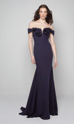 A woman models the Alyce 7062 long formal dress in midnight, featuring an off-the-shoulder neckline with 3D floral accents. She stands against a plain light background. Ideal attire for elegant evening events.