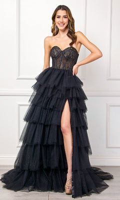 A woman poses indoors against a white paneled wall, wearing the Strapless Ruffled Prom Gown: Amelia Couture 7067—a black evening gown with a sheer bodice, tiered ruffle skirt, and high slit.