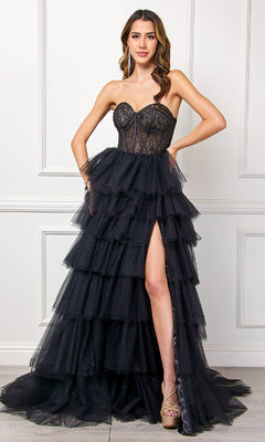 A woman poses against a white paneled wall in the Strapless Ruffled Prom Gown, Amelia Couture 7067, featuring a tiered black skirt, sheer bodice, and high slit.