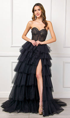 A woman wearing the Amelia Couture 7067 Strapless Ruffled Prom Gown with a sheer bodice and high slit stands against a white paneled wall.