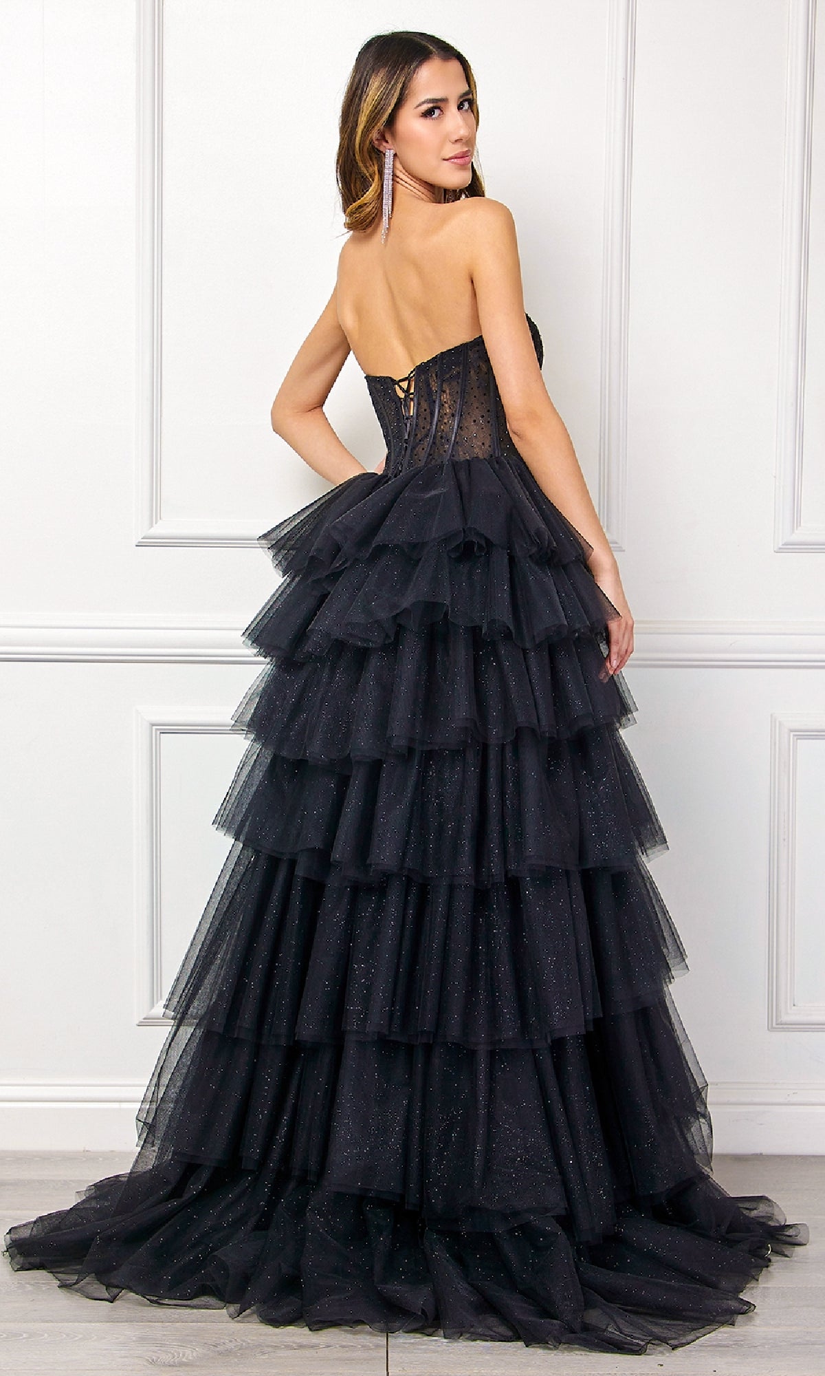 A woman in the Amelia Couture 7067 Strapless Ruffled Prom Gown, featuring a fitted bodice and layered tulle ruffles, stands against a white paneled wall, facing slightly away from the camera.