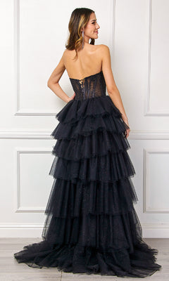 A woman faces away from the camera wearing the Amelia Couture 7067, a strapless ruffled prom gown with a lace-up back and tiered tulle ruffles, set before a white paneled wall.