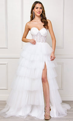A woman in the Strapless Ruffled Prom Gown: Amelia Couture 7067 with a sheer glitter-lace corset bodice, layered tulle skirt, and thigh-high slit stands against a white paneled wall.