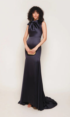 A woman wearing the Alyce 7067 long white sleeveless satin gown with a sheath silhouette and bow neckline holds a small gold clutch, standing against a plain background.