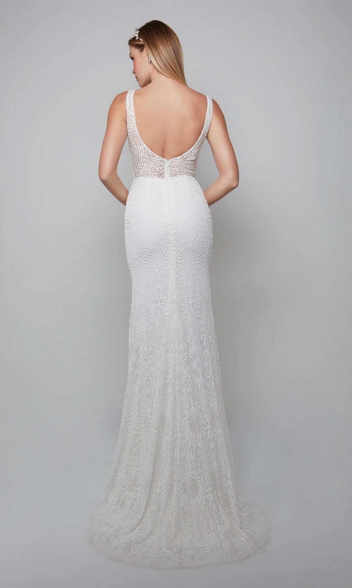 A woman in the Long White Dress: Alyce 7075, a sleeveless white sheath gown with lace appliqués and a long train, stands facing away against a plain gray background.