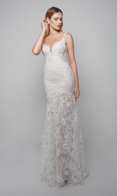 A woman poses against a light gray background wearing the Alyce 7077, a fitted floor-length ivory/lt latte dress in a trumpet silhouette with thin straps, lace appliques, and a deep V neckline.