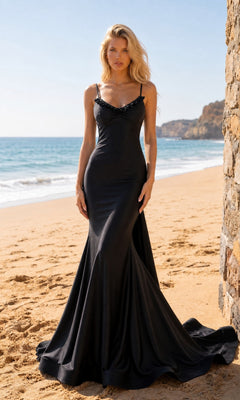A woman wearing the Long Prom Dress: Atria 7082H in black, featuring elegant ruffle detailing and a fitted silhouette, stands on a sandy beach near a stone wall, with ocean waves and rocky cliffs in the background.