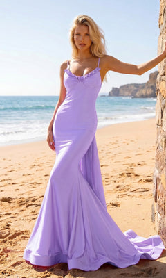 A woman wearing the Atria 7082H long prom dress in lilac, featuring elegant ruffle detailing, stands on a sandy beach near a stone wall with the ocean and cliffs visible in the background.
