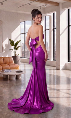 A woman wearing the Atria 7083HF Long Prom Dress—a shiny, floor-length, strapless metallic gown with a large back bow—stands in a modern living room with large windows.