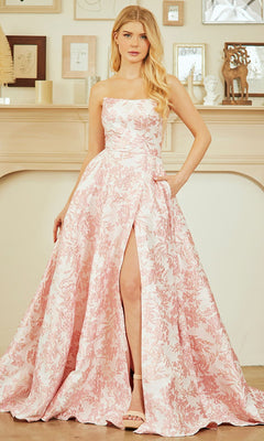 A woman stands indoors in front of a decorated mantel, wearing the Amelia Couture 7084 long prom dress—strapless, floor-length, with pastel floral print and a thigh-high slit.