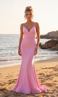 A woman wearing the Atria 7084H long prom dress in light pink stands on a sandy beach near the water, with rocky formations in the background at sunset.