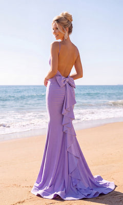 Wearing the Long Prom Dress: Atria 7084H—a backless lavender gown with delicate ruffle details—a woman stands on the sandy beach, facing the ocean beneath a clear sky. The elegant open-back silhouette of the dress beautifully accentuates her figure as she gazes towards the horizon.