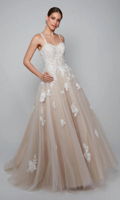A woman wears the Alyce 7084 Long White Dress featuring white floral lace appliqués, thin straps, a fitted bodice, and a graceful silhouette, standing against a plain gray background.