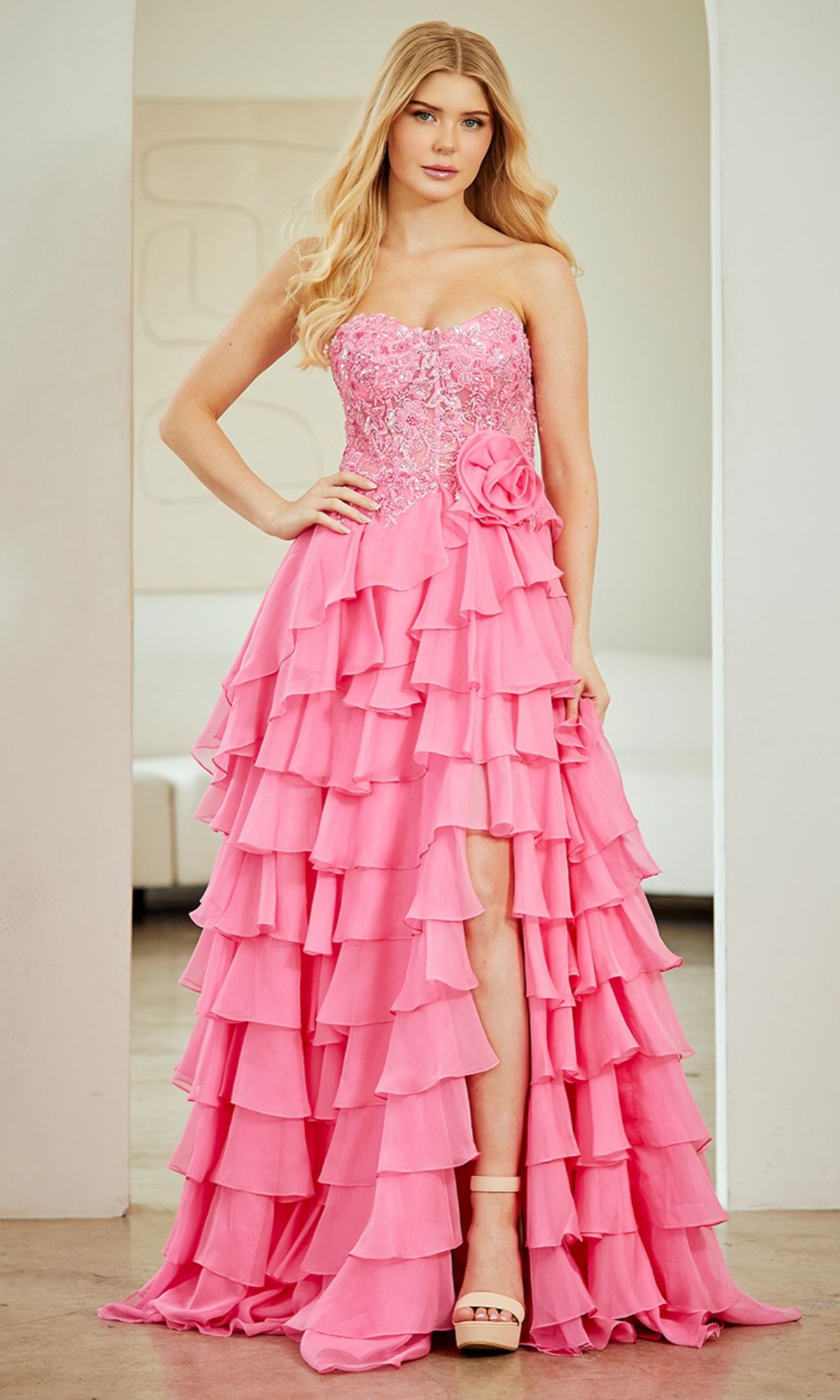 A woman wearing the Strapless Ruffled Prom Gown: Amelia Couture 7087, featuring pink ruffles and a floral waist accent, stands indoors with one hand on her hip.