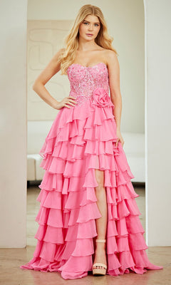 A woman wearing the Strapless Ruffled Prom Gown: Amelia Couture 7087, featuring pink ruffles and a floral waist accent, stands indoors with one hand on her hip.