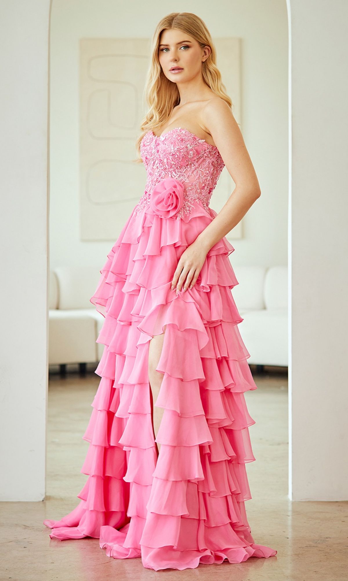A woman wears the Amelia Couture 7087 Strapless Ruffled Prom Gown with layered ruffles and floral details, standing in a modern white room.