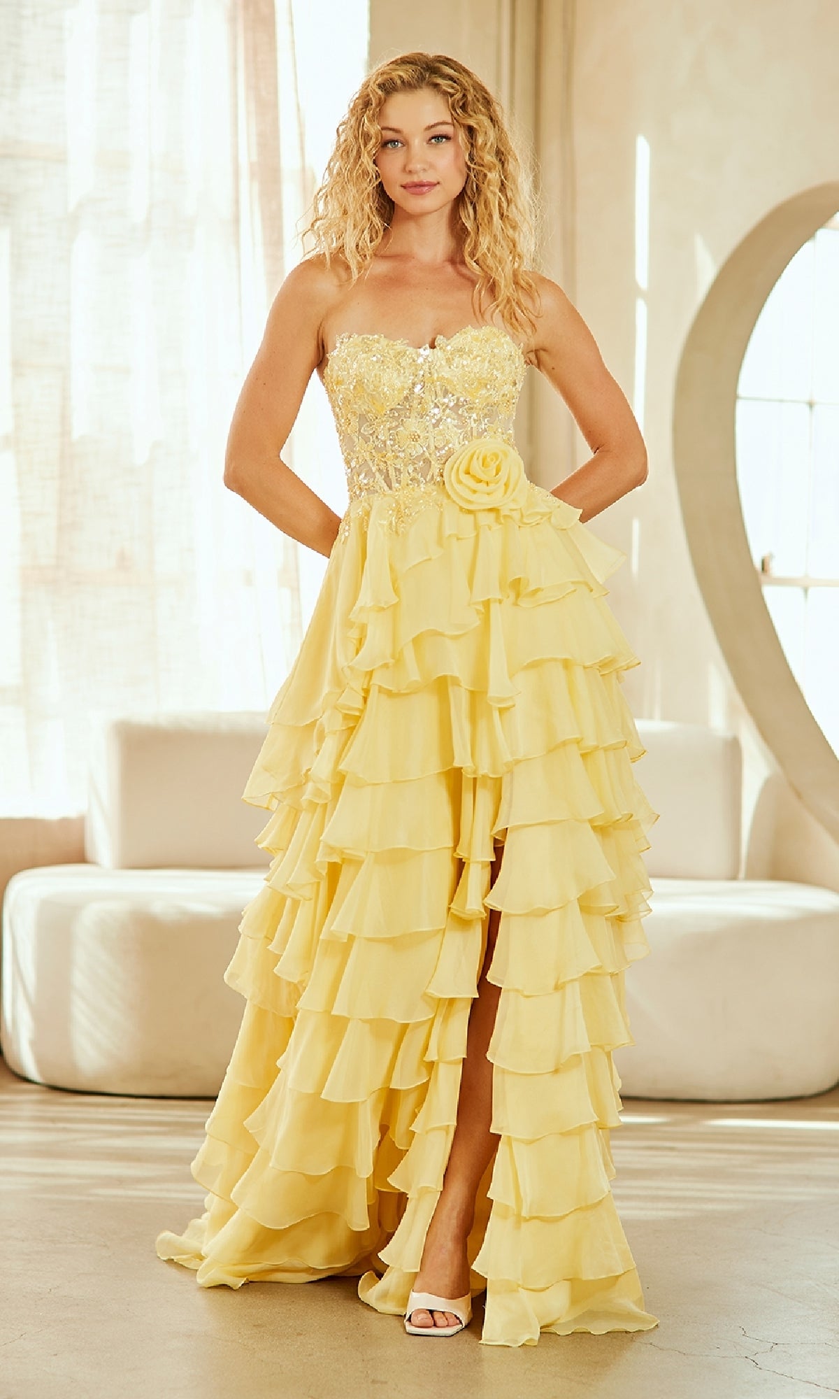 A woman stands indoors in soft light wearing the Amelia Couture 7087, a strapless yellow prom gown with ruffles, a sequined bodice, and floral waist detail.