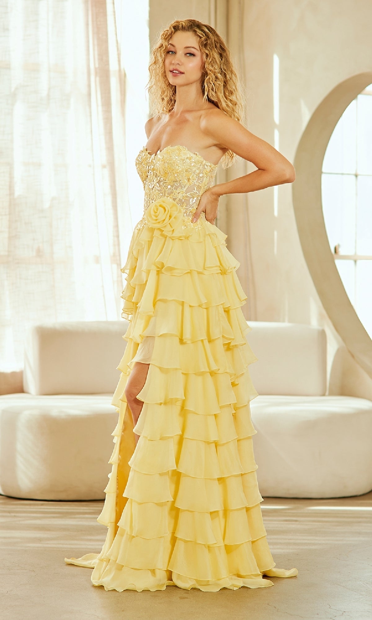 A woman poses indoors in front of a white couch and large window, wearing the Strapless Ruffled Prom Gown: Amelia Couture 7087, a yellow layered dress with floral waist detailing.