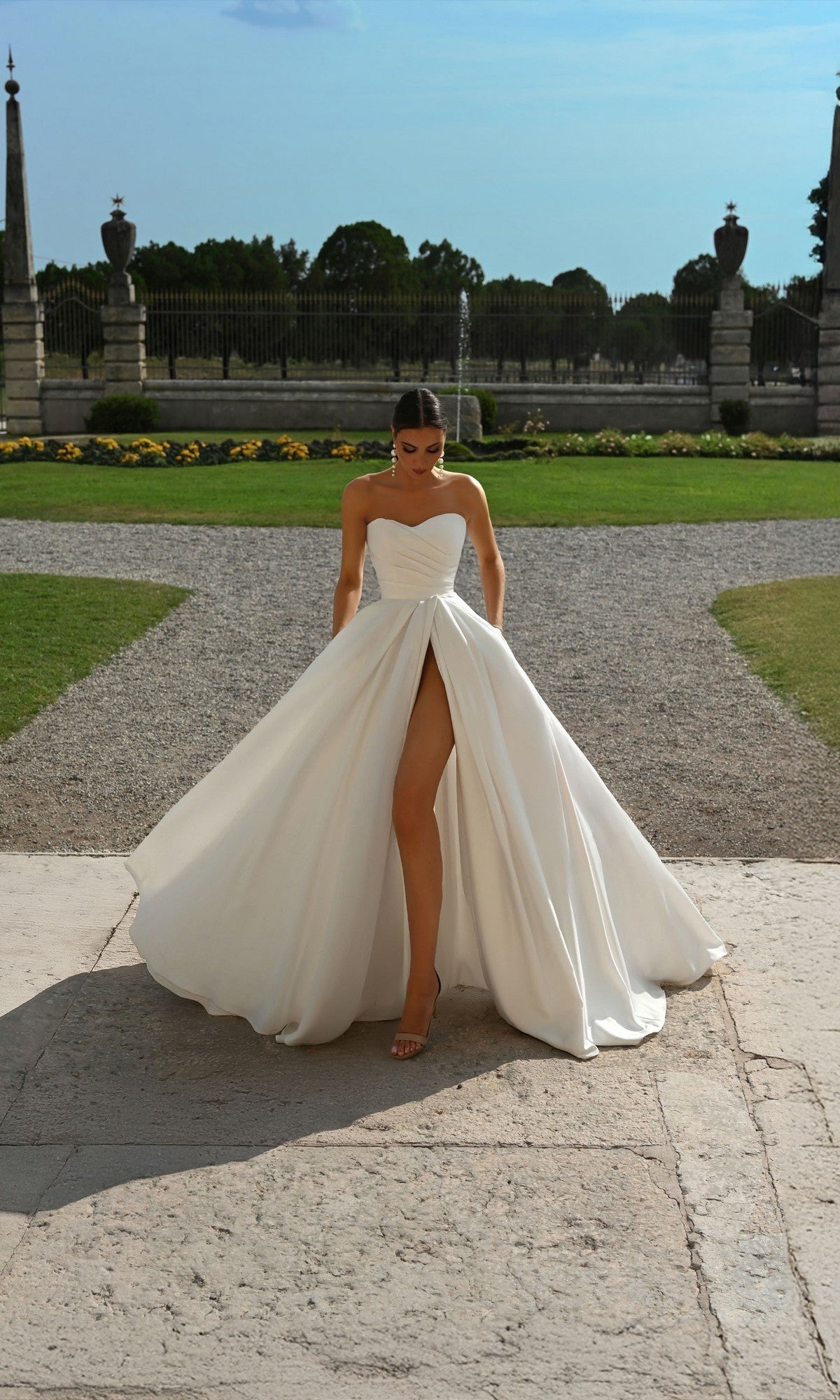 A woman in the Alyce 7088 Long Formal Dress, a strapless satin gown with a high slit, stands outdoors on stone pavement with gardens and a wrought iron fence behind her.