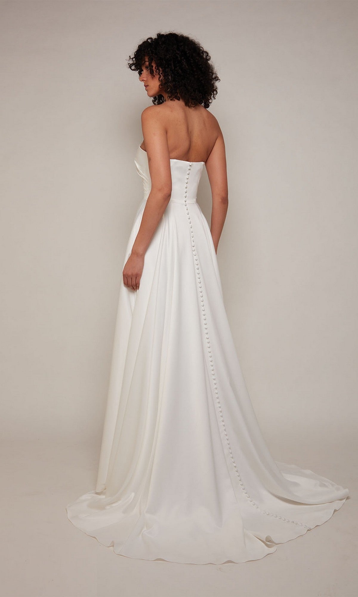 A woman stands facing away in a neutral studio, wearing the Alyce 7088 long formal dress—a strapless satin gown with a long train and button detailing.