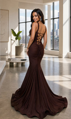 A woman wearing the Atria 7088H Long Prom Dress—a fitted, floor-length dark brown mermaid gown with an adjustable back tie—stands in a modern room featuring large windows and city views.