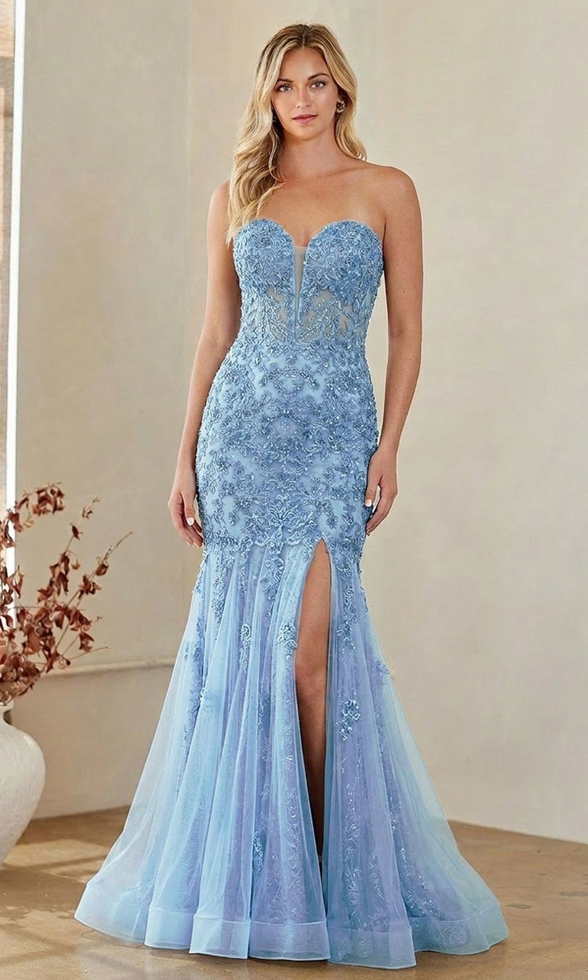 A woman stands indoors wearing the Amelia Couture 7090 Strapless Mermaid Prom Gown in light blue, featuring floral lace details and a high front slit.