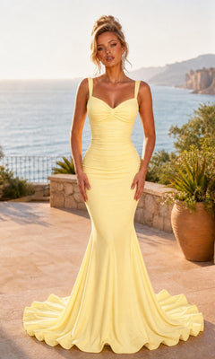 A woman wearing the Atria 7090H Long Prom Dress in soft yellow, featuring a fitted, floor-length design with an elegant lace-up back, stands outdoors on a stone terrace overlooking the ocean and cliffs.