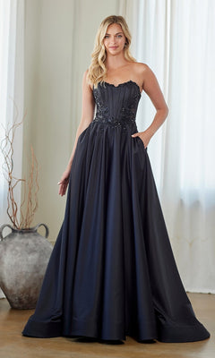 A woman stands indoors wearing the Amelia Couture 7093 long black prom dress, styled with a strapless embroidered corset bodice and a full, elegant A-line skirt.
