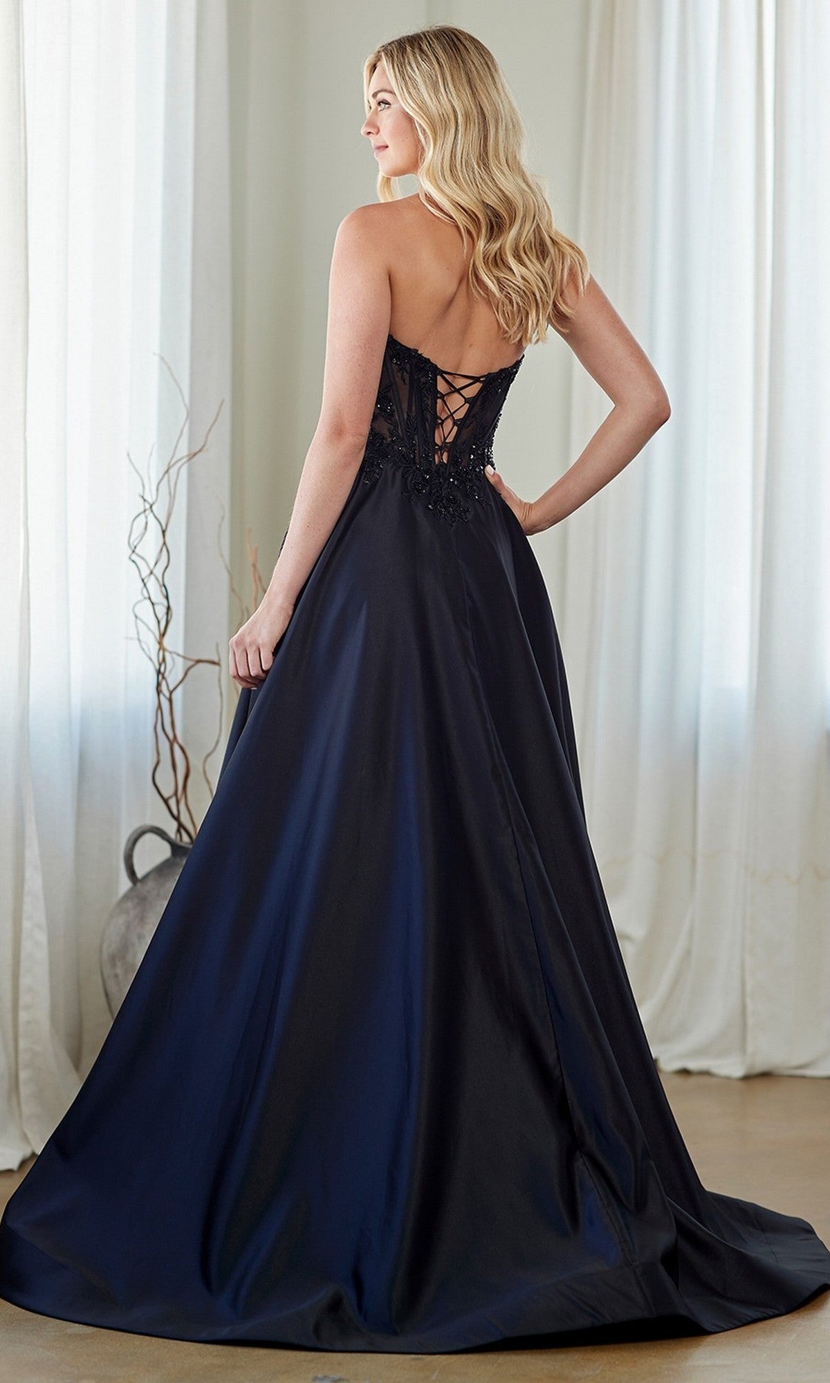 A woman in the Amelia Couture 7093 long prom dress with a strapless embroidered corset bodice and lace-up back stands indoors by white curtains, facing away from the camera.