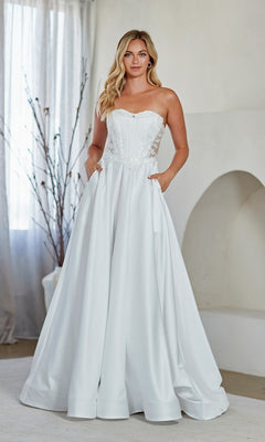 A woman stands indoors wearing the white Amelia Couture 7093 long prom dress, which features a strapless embroidered corset bodice, full A-line skirt, and pockets, set against a neutral background with curtains and decorative branches.
