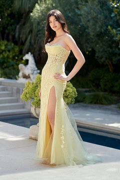 A woman wears a pale yellow, strapless Amelia Couture 7096 prom dress with a lace overlay, sweetheart neckline, and a high side slit.
