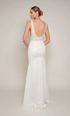 A woman stands with hands behind her back, facing away in the Alyce 7097 Long Formal Dress—a sleeveless gown featuring lace appliqué and a low back, set against a plain background.