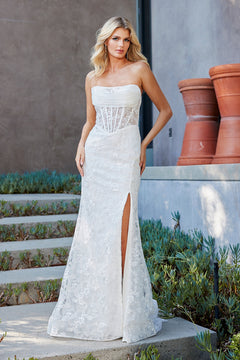 A woman wears a white Amelia Couture 7097 strapless, floor-length lace gown with a corset bodice and a high side slit.