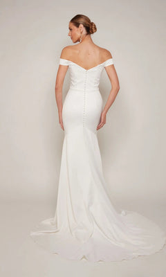 A woman faces away, wearing the Alyce 7098 long formal dress—an off-the-shoulder white gown with back button details and a long train. Her hair is styled in a sleek low bun.