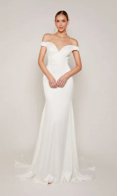 A woman stands before a light background, wearing the Alyce 7098—a white, off-the-shoulder long formal dress featuring a sweetheart neckline, fitted silhouette, and flared floor-length hem.