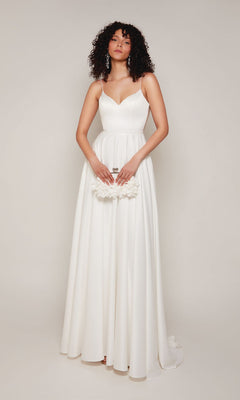 A woman in the Alyce 7099 long white dress with beaded spaghetti straps holds a small white clutch, standing against a plain light background.