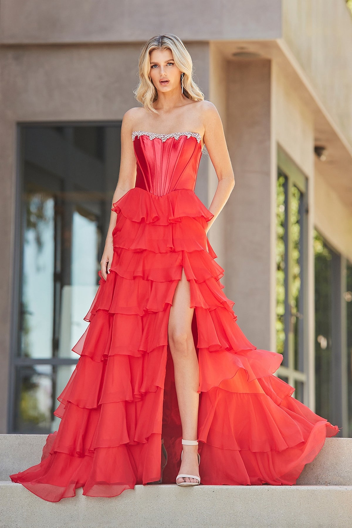 A model wearing the Amelia Couture 7099, a red strapless ruffled corset dress with a layered skirt and a high leg slit.