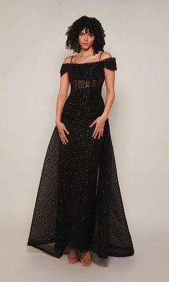 A person stands against a plain background wearing the Alyce 7101—a long, black formal dress with thin straps, off-the-shoulder style, and sparkling glitter details.